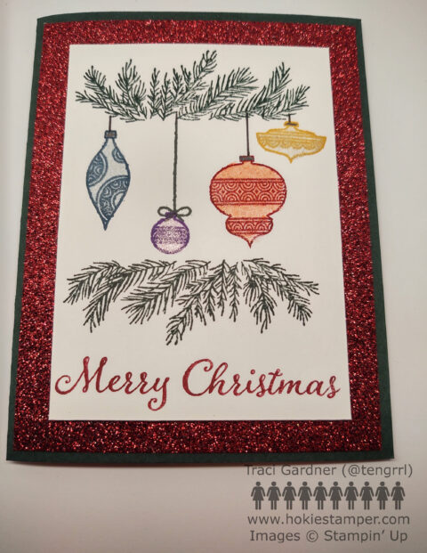 Christmas card showing pine branches framing four hanging ornaments in blue, purple, red, and yellow. The sentiment is Merry Christmas. The pine branch layer is mounted on red glitter paper.