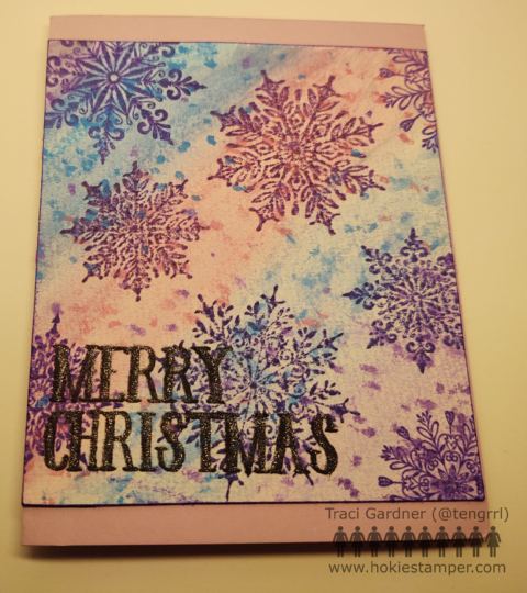 Christmas card showing purple snowflakes on a pink, blue and purple watercolor background. The sentiment Merry Christmas is stamped in black on the lower left corner. The sentiment sparkles from a layer of glitter.