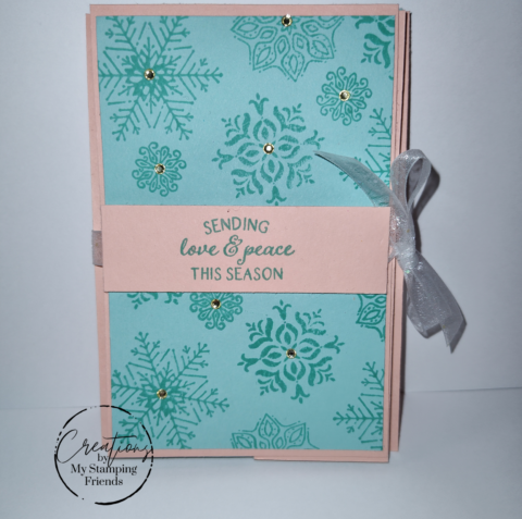 Front of a pink and blue greeting card, with a variety of blue snowflakes in the background and the sentiment, Sending love and peace this season. The card ties closed with white ribbon on the right side.